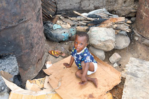 A little african boy is sitting between rusty metal barrels used for fish smoking