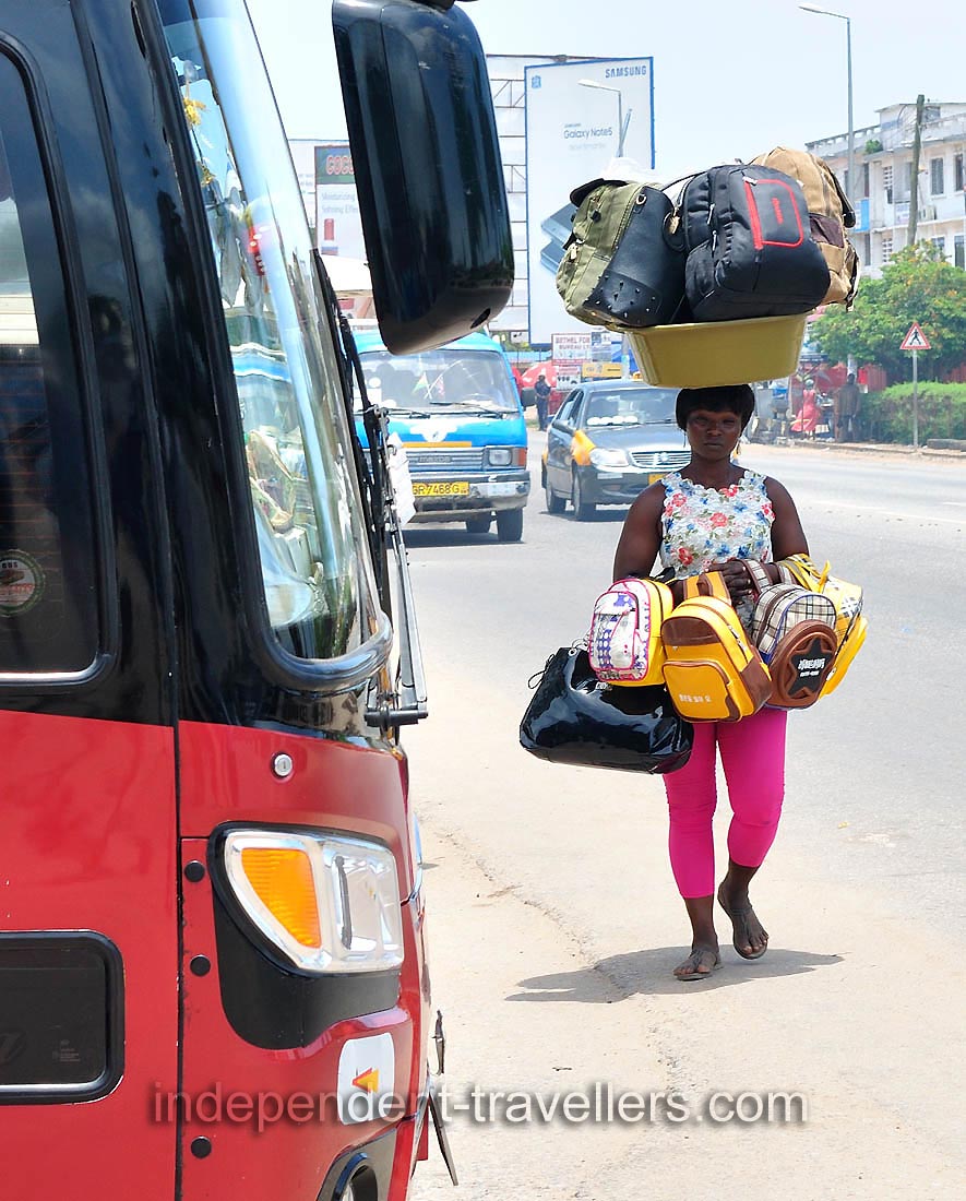 A female vendor is carrying huge travel bags on the head and in the hands