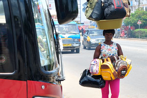 A female vendor is carrying huge travel bags on the head and in the hands