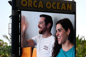 A spanish couple is smiling from a large outdoor screen before the orca show