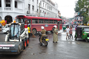 A Bobcat road roller, a red bus and an autorickshaw are on the intersection